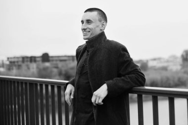 Yakışıklı delikanlı paltolu. Moda iyi giyimli erkek gülümseme şık ceket. Çocuk sonbahar açık. Mükemmel saç modeli olan zarif adam. Köprüde poz. Moda siyah ve beyaz. Adam gülümseme