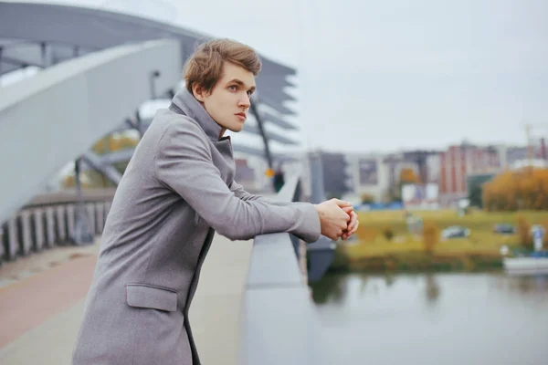 Young handsome man in coat. Fashionable well dressed boy smile in stylish coat. Boy outdoor at autumn. Elegant man with perfect hairstyle. Posing on the bridge. Fashion