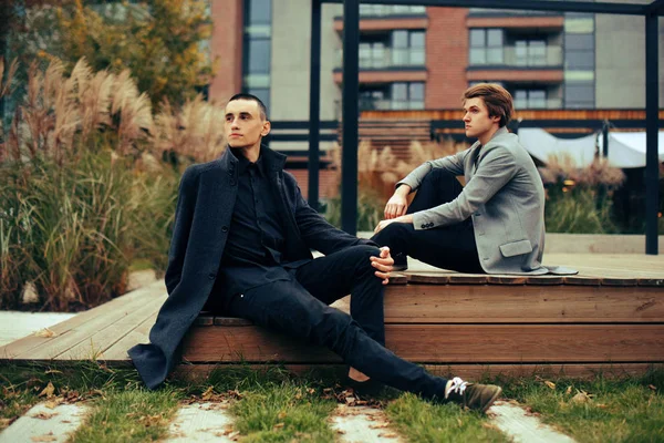 Two friends sit in the park on a bench. Friendship. Two fashion men in coats .