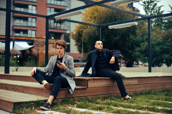 Two friends sit in the park on a bench. Friendship. Two fashion men in coats  . Man vape, his friend uses the phone