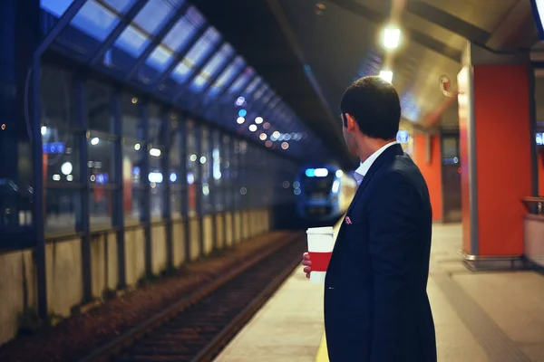 Arka planda tren hareket ile tren istasyonunda kahve olan bir adam duruyor. Bir tren istasyonu platformu üzerinde duran bir beyefendi. iş seyahat, bir tren bekleyen iş adamı.