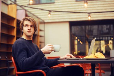 Adam bir restoranda terasta kahve içme. Serbest meslek ile bir dizüstü bilgisayar, açık kruvasan kahvaltı vardır. İşadamı görünüyor ve rüyalar. Dizüstü olan adam. Kablosuz kulaklık. 