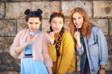Üç güzel kız model lolipop yemek. Kız arkadaşları değişik saç modelleri ve stilleri ile. Kız öpücük, wriggles göndermek, gülmek, eğlenmek. Güneş gözlüğü ve giyimleri dışarıda kızlar. Şirin 