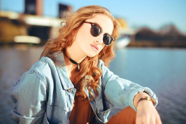 İskeleye Nehri yakınında oturan güzel kız. Hipster modeli muhteşem cerly saçlı güneş gözlüğü. Kız Kot ceket içinde mesafe içine güneşli gün görünüyor. Yaz saati. Gerdanlık kız