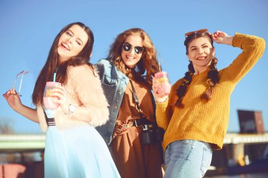 Üç kızlar eğleniyor gökyüzü karşı Nehri yakınında. Güneş gözlüğü moda kızlar. Atlama, sevinirler, içecekler için gülümse kafayı iyi eğlenceler. Renk giyinmiş arkadaşlar harcamak yaz vibes, zaman birlikte.