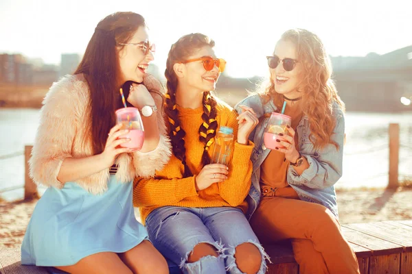 Three girls sit on the beach , near the river, talk, gossip, joke, laugh, smile at womans day. Girls on a hot day drink lemonade. Hipster model wearing sunglasses at summer day. Summer vibes.