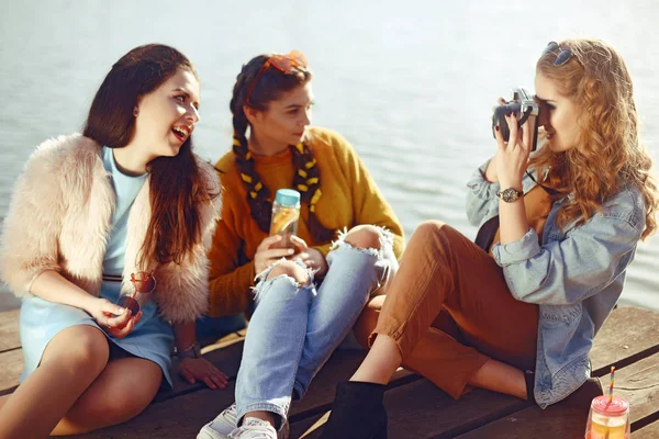 The blonde girl takes pictures of her brunette girlfriends on the old camera. They send her kisses, smile, laugh. Girl with drinks and sunglasses have fun. Girls on the dock/pier on the back of yachts