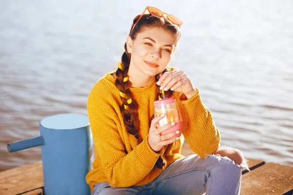 The girl at hot sunny day drink lemonad. Summer vibes. Girl with fashion yellow sunglasses and dressed in yellow. Beautiful girl sitting on the pier near the river. Beauty model with brunette hair.