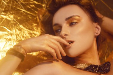 Altın Moda Makyajı, Sanat Güzelliği Yüzü ve Dudaklar Altın Brokar ile Makyaj. Moda altın tenli kadın portresi. Altın mücevherler, mücevherler, aksesuarlar, küpeler. Altın gölgeler. Kız yalan söylüyor.