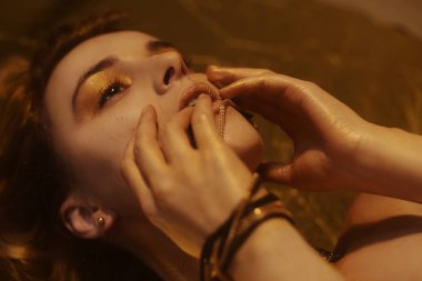 Altın Moda Makyajı, Sanat Güzelliği Yüzü ve Dudaklar Altın Brokar ile Makyaj. Moda altın tenli kadın portresi. Altın mücevherler, mücevherler, aksesuarlar, küpeler. Altın gölgeler. Kız yalan söylüyor.