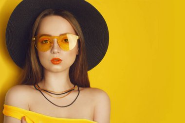 Turuncu profesyonel görünüm aksesuarları ile Güzellik Modeli. Uzun, düz saçlı moda kadını. Trend makyaj. Turuncu arka plan. Büyük siyah şapkalı ve sarı güneş gözlüklüğü olan bir kız. Boş kopyalama alanı