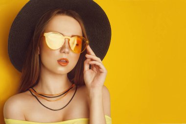 Turuncu profesyonel görünüm aksesuarları ile Güzellik Modeli. Uzun, düz saçlı moda kadını. Trend makyaj. Turuncu arka plan. Büyük siyah şapkalı ve sarı güneş gözlüklüğü olan bir kız. Boş kopyalama alanı