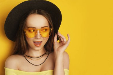 Turuncu profesyonel görünüm, aksesuarlar ile Güzellik Modeli. Uzun saçlı moda kadını. Trend makyaj. Turuncu arka plan. Büyük siyah şapkalı ve sarı güneş gözlüklüğü olan bir kız. Kız heyecanlı görünüyor.