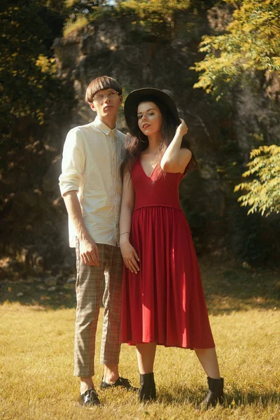 美丽的时尚时尚夫妇在森林公园 女孩在美丽的大帽子和红色的长裙 一个男人穿着时髦的白衬衫和眼镜 夏天一对非常时髦的人 — 图库照片
