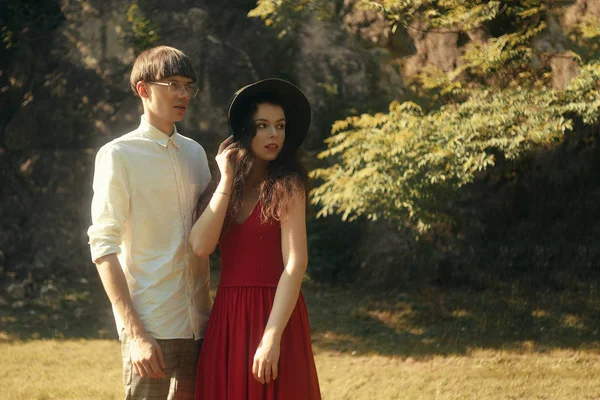 美丽的时尚时尚夫妇在森林公园 女孩在美丽的大帽子和红色的长裙 一个男人穿着时髦的白衬衫和眼镜 夏天一对非常时髦的人 — 图库照片