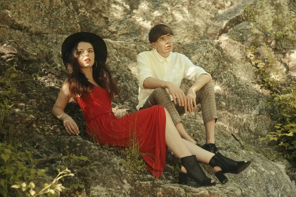 时髦的夫妇坐在岩石上 时髦的女人穿着时髦的红色长裙 山中的男人和女人戴着帽子的布鲁内特一个留着白衬衫和眼镜的男人 — 图库照片