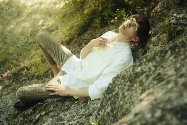 Fashionable hipster guy lying on the rocks. Hipster man with a fashionable haircut. Man in cool glasses and white shirt. Man in the mountains.