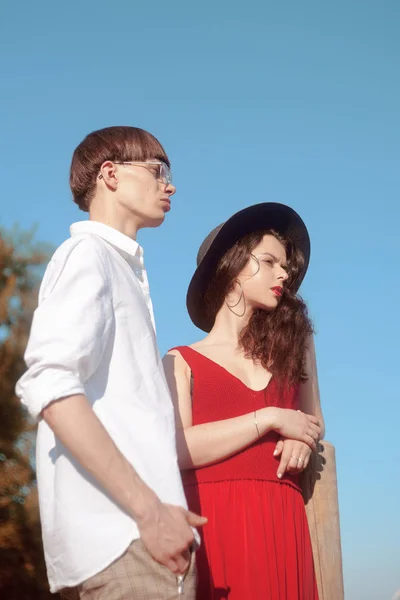 Beautiful fashionable couple on the beach near the river. Fashionable hipsters. Girl in a red dress and black big hat. A man with glasses and a white shirt. Model pouse. Enjoy a sunny summer day.