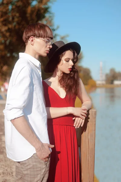 美丽的时尚夫妇在河边的海滩上 时尚时尚 女孩穿红色连衣裙 戴着黑色大帽子 戴眼镜和白衬衫的男人模型粪便 享受阳光明媚的夏日 — 图库照片