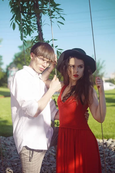 Beautiful fashionable couple against the sky and the beach . Fashionable hipsters. Girl in a red dress and black big hat. A man with glasses and a white shirt. Model pouse at holiday, relaxation.