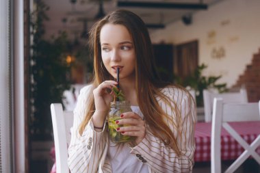 Nehir kenarındaki gemideki bir restoranda bulunan kız mojito içiyor. Esmer gülümsüyor ve bardak kavanozda mojito içiyor. Ceketli uzun saçlı kız, sıcak bir günde mola. İşten sonra kız, öğrenci.