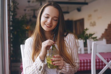 Nehir kenarındaki gemideki bir restoranda bulunan kız mojito içiyor. Esmer gülümsüyor ve bardak kavanozda mojito içiyor. Ceketli uzun saçlı kız, sıcak bir günde mola. İşten sonra kız, öğrenci.