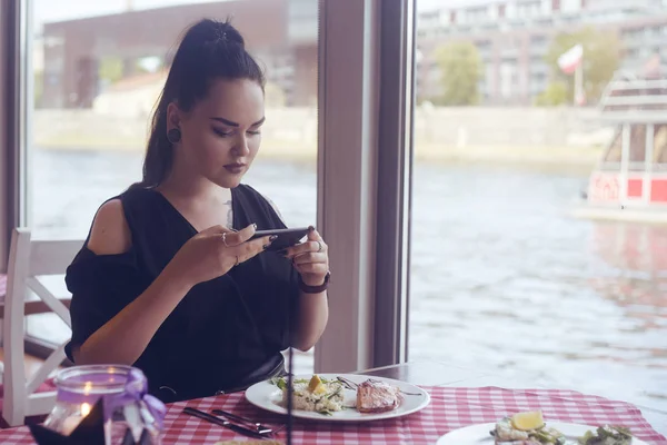 Girl food blogger takes a picture on the phone from social networks. Woman shooting food in a restaurant. Brunette makes a photo of salmon and risotto, dish. National food, tourist smile. Roaming.