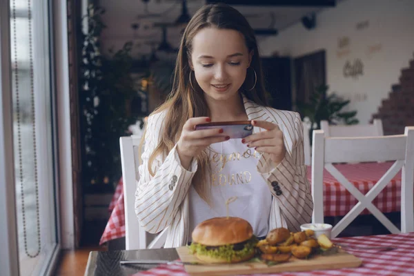 长头发的女孩坐在餐厅里 女孩食品博主在手机上从社交网络上拍照 女人在餐馆里吃菜制作汉堡和炸土豆的照片 — 图库照片