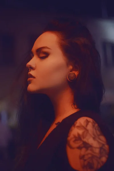 The girl in the tattoo, with piercing in the nose and tunnels in the ears. Beautiful girl, night portrait. The girl in neon lighting. Beautiful unconventional brunette. Girl with a professional make-up.