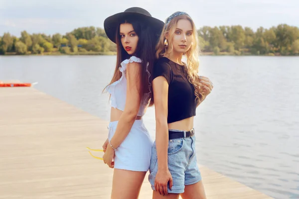 Twee modieuze meisjes, vrienden, staan op de pier in de buurt van de rivier — Stockfoto