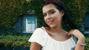 Güzel bir esmer, yaprakların arasında bir evin arka planında poz veriyor ve gülümsüyor. İspanya 'da tatiller. Küpeli çemberli bir kız. Kameraya bakıyor, gülümsüyor.