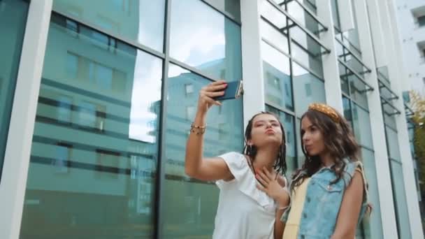 两个女朋友 姐妹走在新大楼 新办公室 新城市的附近 女孩们有乐趣 游客们走着笑着 在电话上给自己拍照 — 图库视频影像