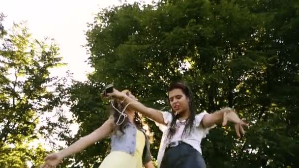 两个女朋友成双成对地跳舞 听音乐 女孩们通过耳机听音乐 唱歌和跳舞 女孩们在草地上跳舞 — 图库视频影像