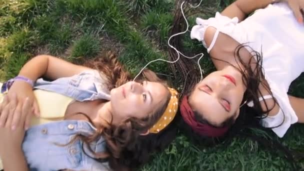 二人のガールフレンド 姉妹が草の上に横たわり ヘッドフォンで音楽を聴いています 二つのブルネットは 公園の草の上に横たわって リラックスして — ストック動画