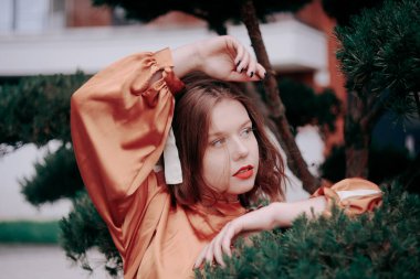 Ahşap, doğa, yeşillik ve ladin arka planında şık, güzel bir model. Kısa saçlı ve kahverengi elbiseli bir kız. Sarışın bir ağaçta yatıyor, manken şık bir şekilde poz veriyor..