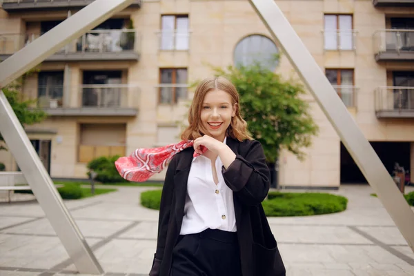 A girl in a business suit with a red scarf on her head against the background of a building, turns to the camera and laughs. Fashion photo of a blonde model poses, in a suit and with a red scarf.