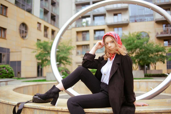 建物を背景に赤いスカーフを頭にしたビジネススーツの少女が噴水の上に座って休んでいる ブロンドモデルのファッション写真 スーツ姿のモデル — ストック写真