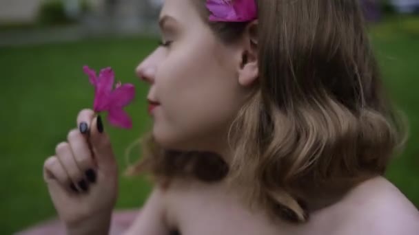 花の髪を持つ豪華な女の子の肖像画 ピンクの花の後ろに対するブロンド ファッショナブルな色合いの夏の写真 短い散髪で美しい化粧で妖精の庭で美しい — ストック動画