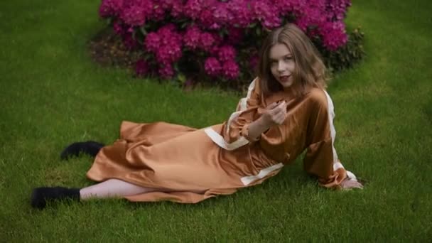 一个模特穿着一件棕色的长裙 坐在花园的草地上 背靠着一丛丛烂花 与自然 美丽的女孩在一起 微笑着 金发碧眼 — 图库视频影像