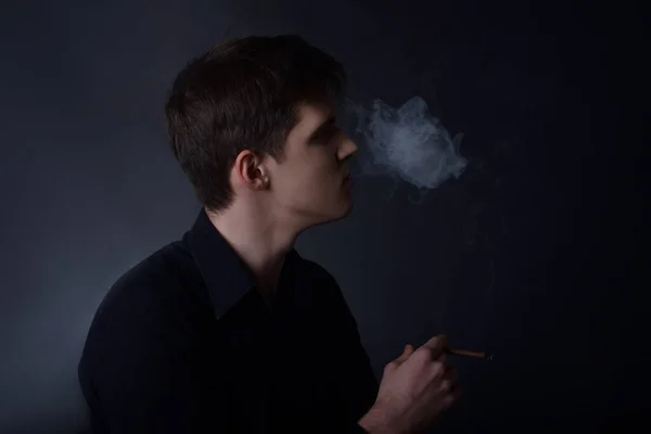 A man in a black shirt in the studio on a black background smokes a cigar, cigarettes, exhales smoke