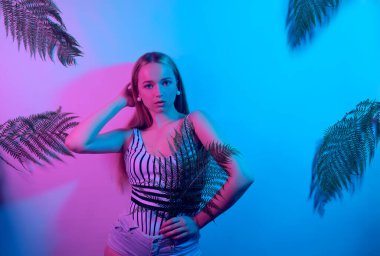 Neon pozlar veren bir model. Mayo giymiş bir kızın moda fotoğrafı ve pembe-mavi ışık altında şortu. Tropikal yaprakların arka planında bir kız yaz fotoğrafı. Güzel sarışın.
