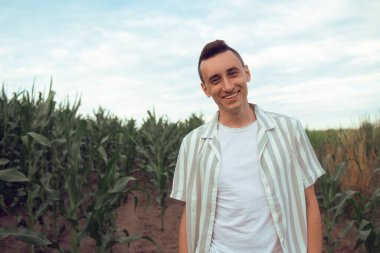 Modaya uygun bir adam mısır tarlasında yürüyor. Şehir dışında güneşli bir gün. Yaprakların ve günbatımının arkasındaki adam gülümsüyor.