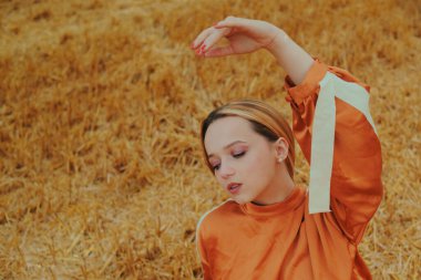 Uzun elbiseli muhteşem bir kız buğday tarlasında oturuyor. Şık bir fotoğraf için sarışın pozu. Kadın ellerini çok güzel oynatıyor..