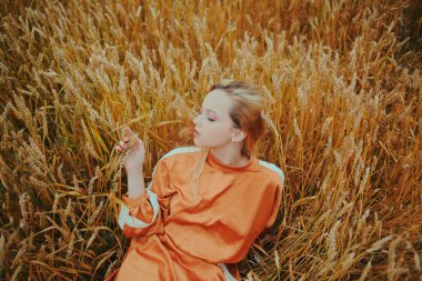 Zarif bir model buğday tarlasında uzanıyor ve doğanın tadını çıkarıyor. Doğal ürünler konsepti, doğayla bütünlük, moda. Uzun saçlı güzel sarışın kız.