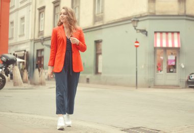 Şehrin eski sokağının önünde poz veren şık hippi kız. Modayı takip eden genç bir kadın model. Sokak modası konsepti, kırmızı ceket giymek. Turist gülümsemesi, güneşli bir gün. Uzun kıvırcık saçlı zarif bir kız.