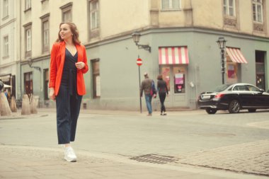 Şehrin eski sokağının önünde poz veren şık hippi kız. Modayı takip eden genç bir kadın model. Sokak modası konsepti, kırmızı ceket giymek. Turist gülümsemesi, güneşli bir gün. Uzun kıvırcık saçlı zarif bir kız.
