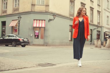 Şehrin eski sokağının önünde poz veren şık hippi kız. Modayı takip eden genç bir kadın model. Sokak modası konsepti, kırmızı ceket giymek. Turist gülümsemesi, güneşli bir gün. Uzun kıvırcık saçlı zarif bir kız.