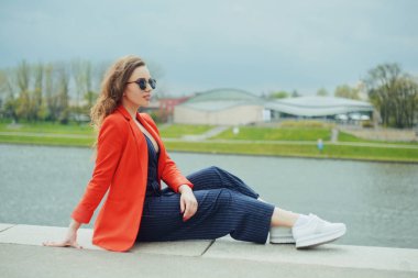 Riverside 'da güneş gözlüğü takan güzel bir kız. Kız iskelede oturuyor ve nehri seyrediyor. Güneşli havada güzel genç kızlar. Moda modeli seyahati. Kırmızı ceketli, uzun saçlı kız..