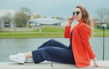 Riverside 'da güneş gözlüğü takan güzel bir kız. Kız iskelede oturuyor ve nehri seyrediyor. Güneşli havada güzel genç kızlar. Moda modeli seyahati. Kırmızı ceketli, uzun saçlı kız..