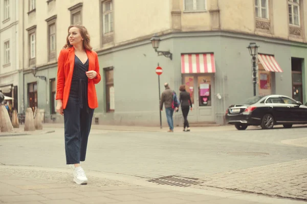 Şehrin eski sokağının önünde poz veren şık hippi kız. Modayı takip eden genç bir kadın model. Sokak modası konsepti, kırmızı ceket giymek. Turist gülümsemesi, güneşli bir gün. Uzun kıvırcık saçlı zarif bir kız.